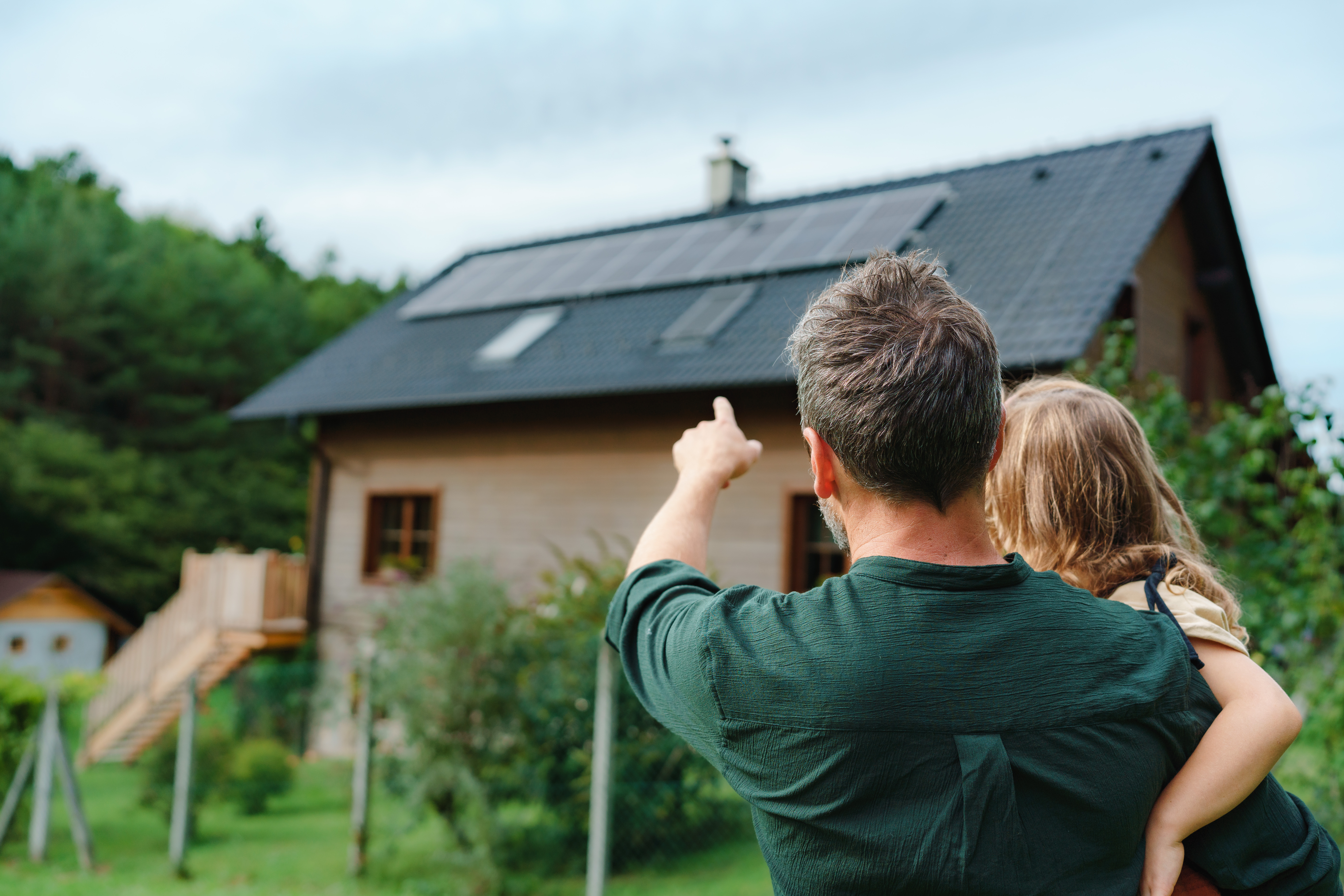 Rear view of dad holding her little girl in arms and showing at their house with solar panels.